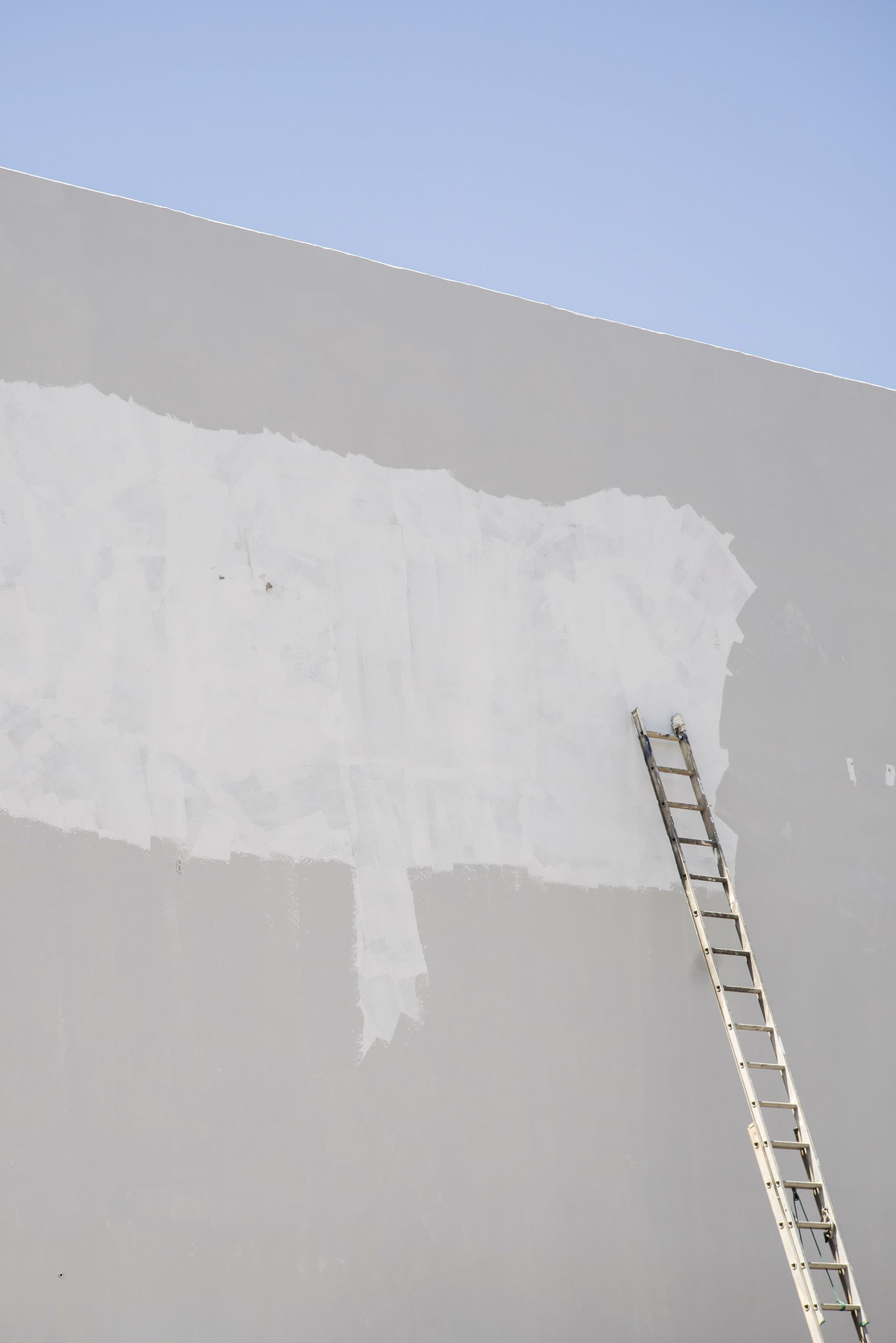 An extension ladder leaning against a partially painted gray wall under a clear blue sky.