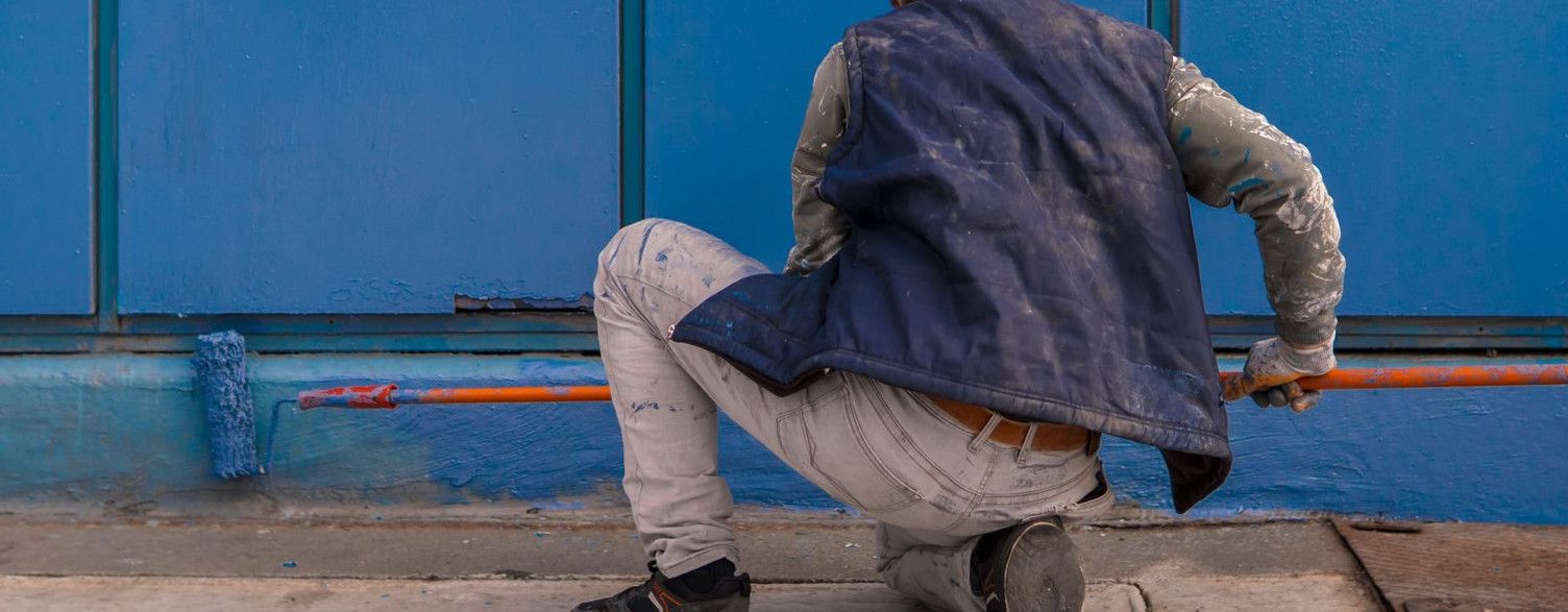 A construction worker paints a large blue wall outside, adding color and life.